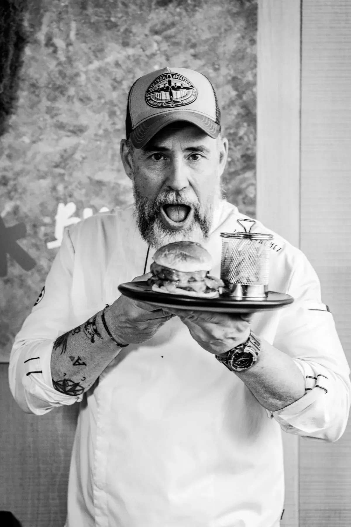 Un chef con barba y gorra de béisbol sostiene un plato con una hamburguesa y papas fritas