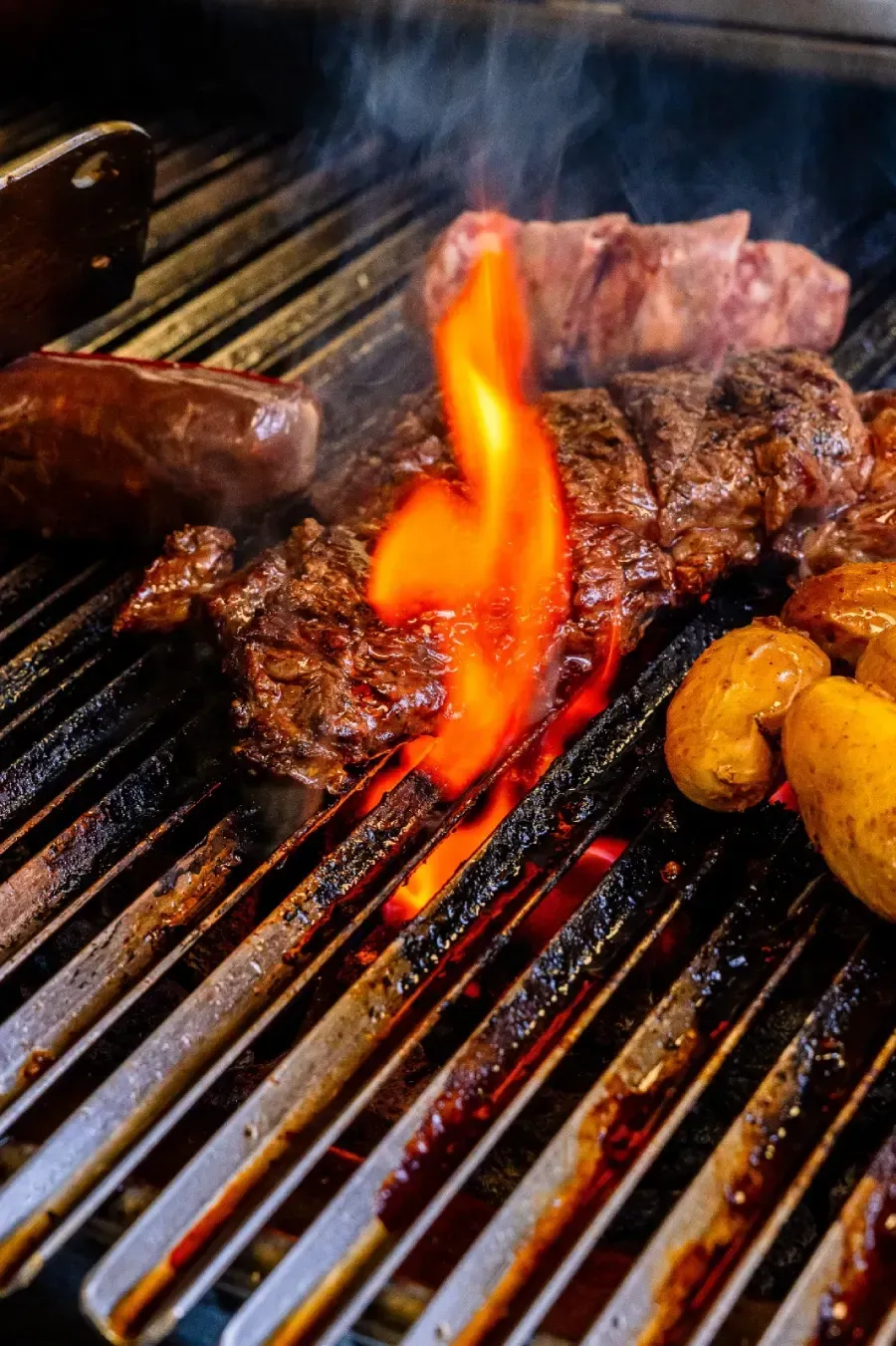 Un trozo de bistec y papas pequeñas asándose en una parrilla de barbacoa al aire libre, caliente y con llamas.