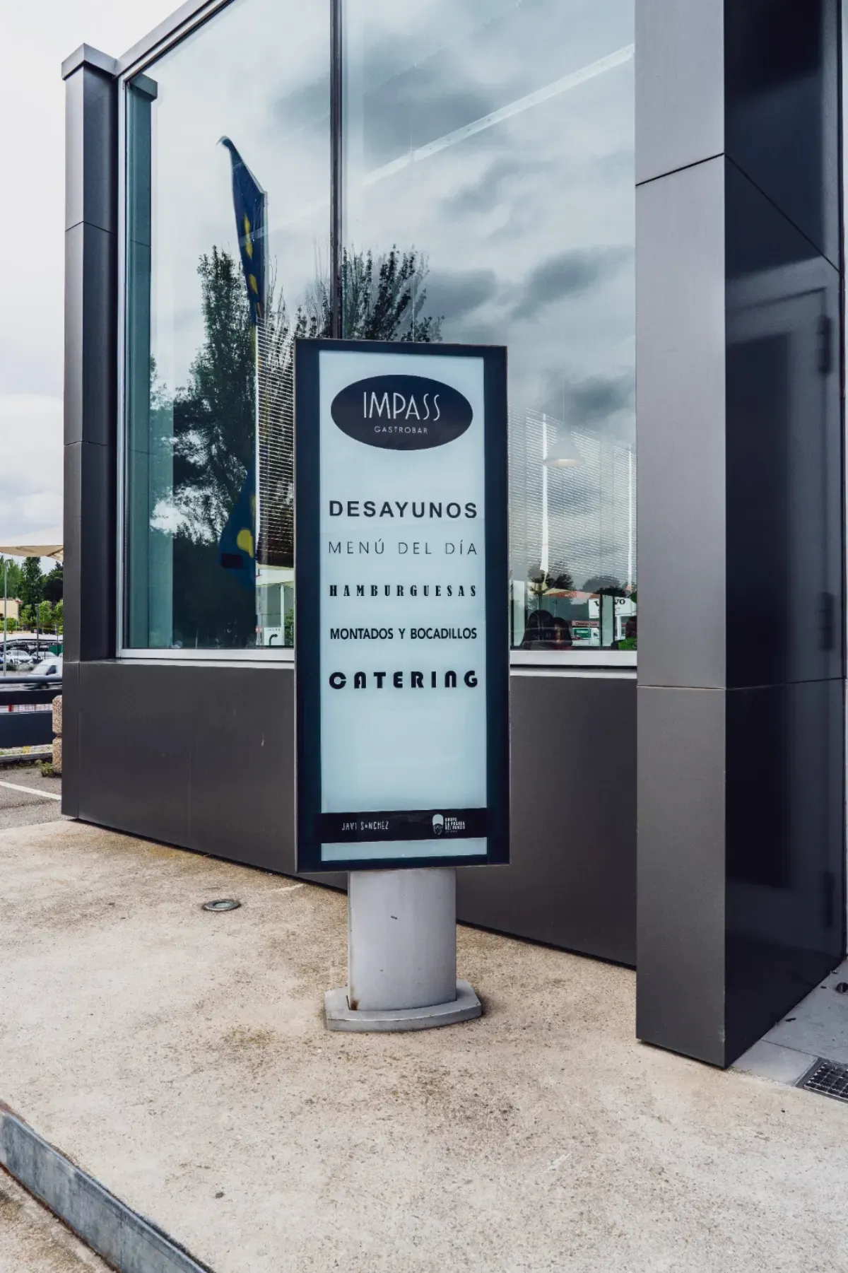 Un cartel informativo vertical se alza sobre una acera de hormigón frente a un moderno edificio con fachada de cristal.