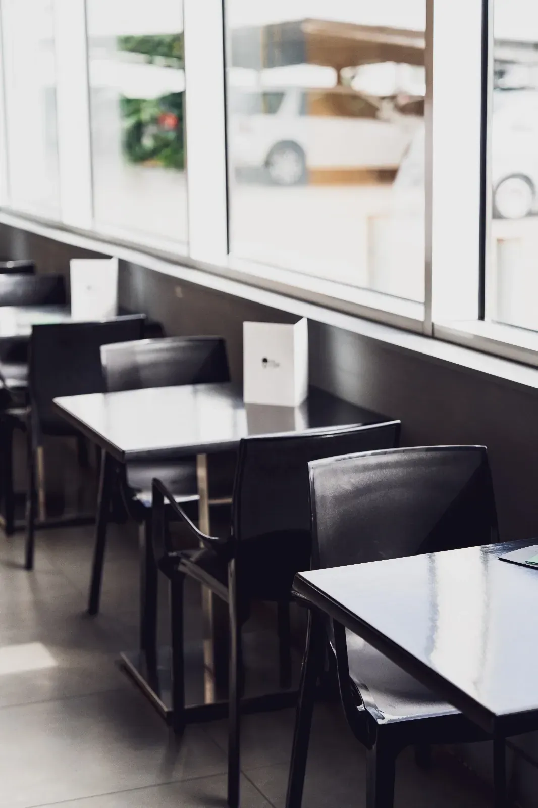Mesas y sillas negras vacías dispuestas en fila dentro de una habitación minimalista y muy iluminada con grandes ventanales.