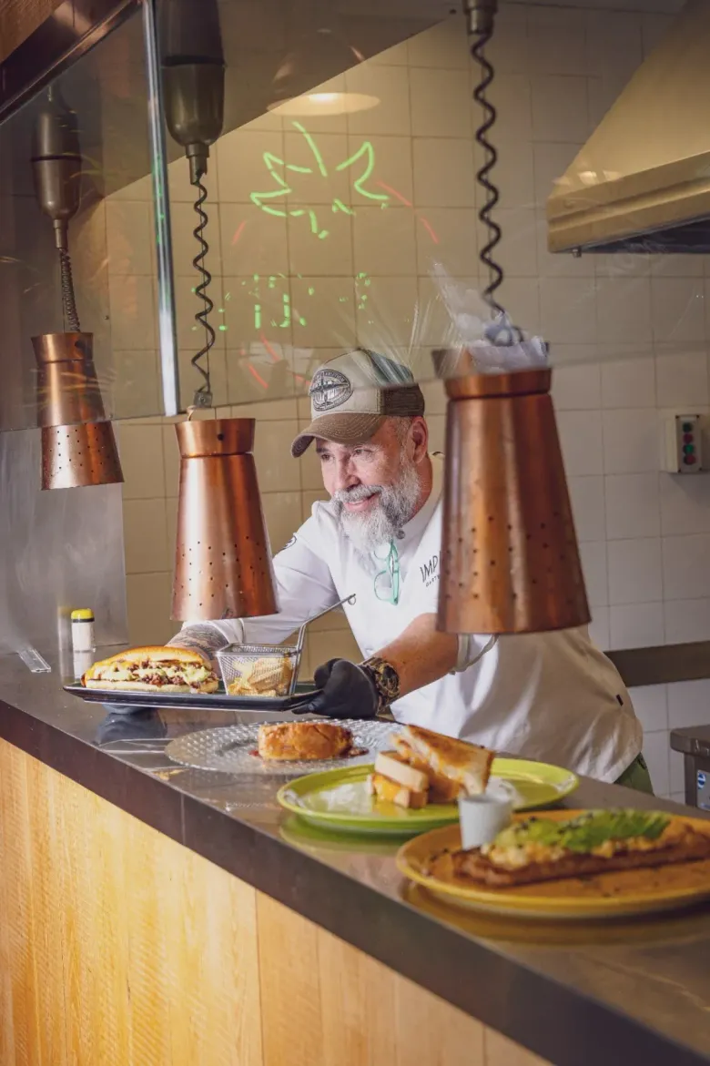 Un chef sonriente, con barba y gorra, sirve sándwiches recién preparados en platos sobre un mostrador