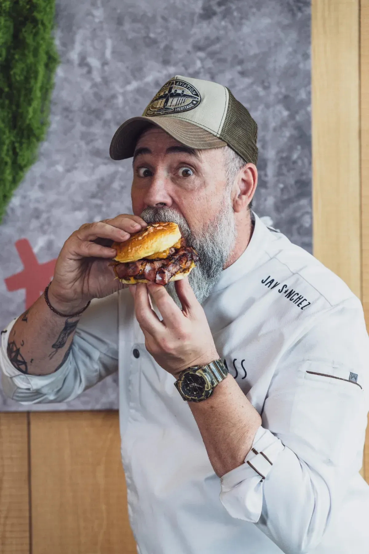 Un chef barbudo, con uniforme blanco y gorra estampada, se lleva una hamburguesa a la boca con una expresión juguetona
