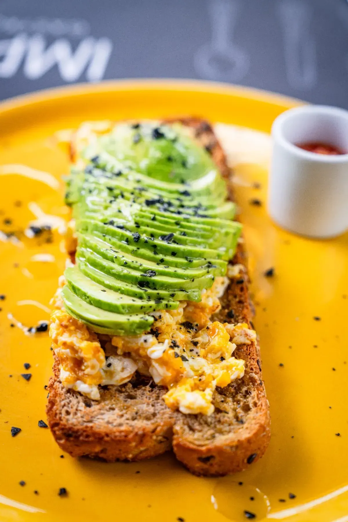 Tostada de aguacate cubierta con huevos revueltos y condimentos en un plato amarillo con una pequeña porción de salsa.