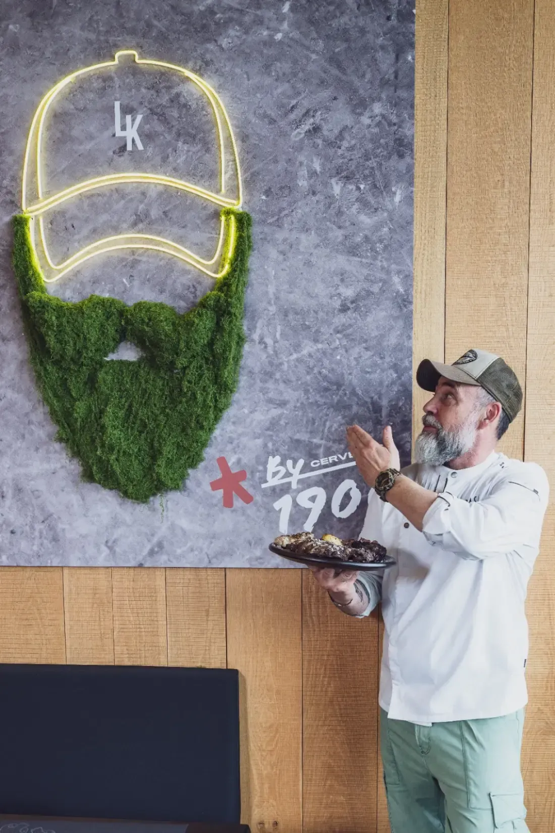 Un hombre barbudo con gorra de béisbol señala una obra de arte en la pared que muestra el contorno de un sombrero de neó