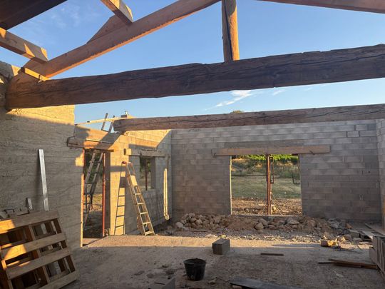 Maison en parpaings partiellement construite, avec des poutres de toit et des ouvertures dans les murs.