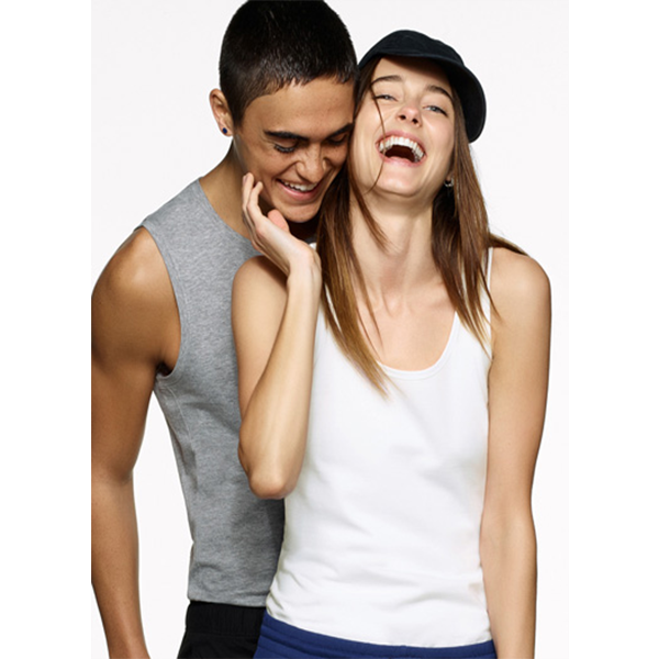 Mann im grauen Tanktop lächelt hinter lachender Frau im weißen Tanktop und schwarzem Hut.