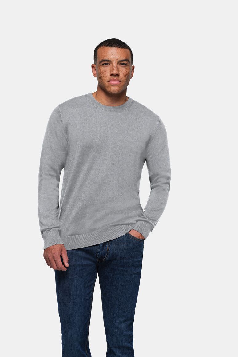 Mann in grauem Pullover und Jeans, Hand in der Tasche, steht vor weißem Hintergrund.