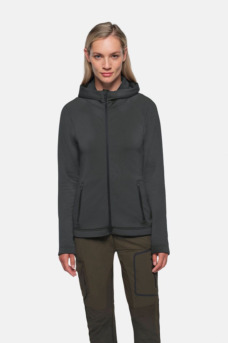 Frau in grauer Kapuzenjacke und grüner Hose, vor hellgrauem Hintergrund stehend.