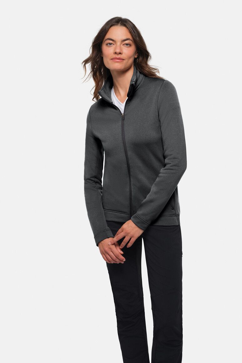 Frau in grauer Jacke mit Reißverschluss und schwarzer Hose, steht im Innenbereich und blickt nach vorne.