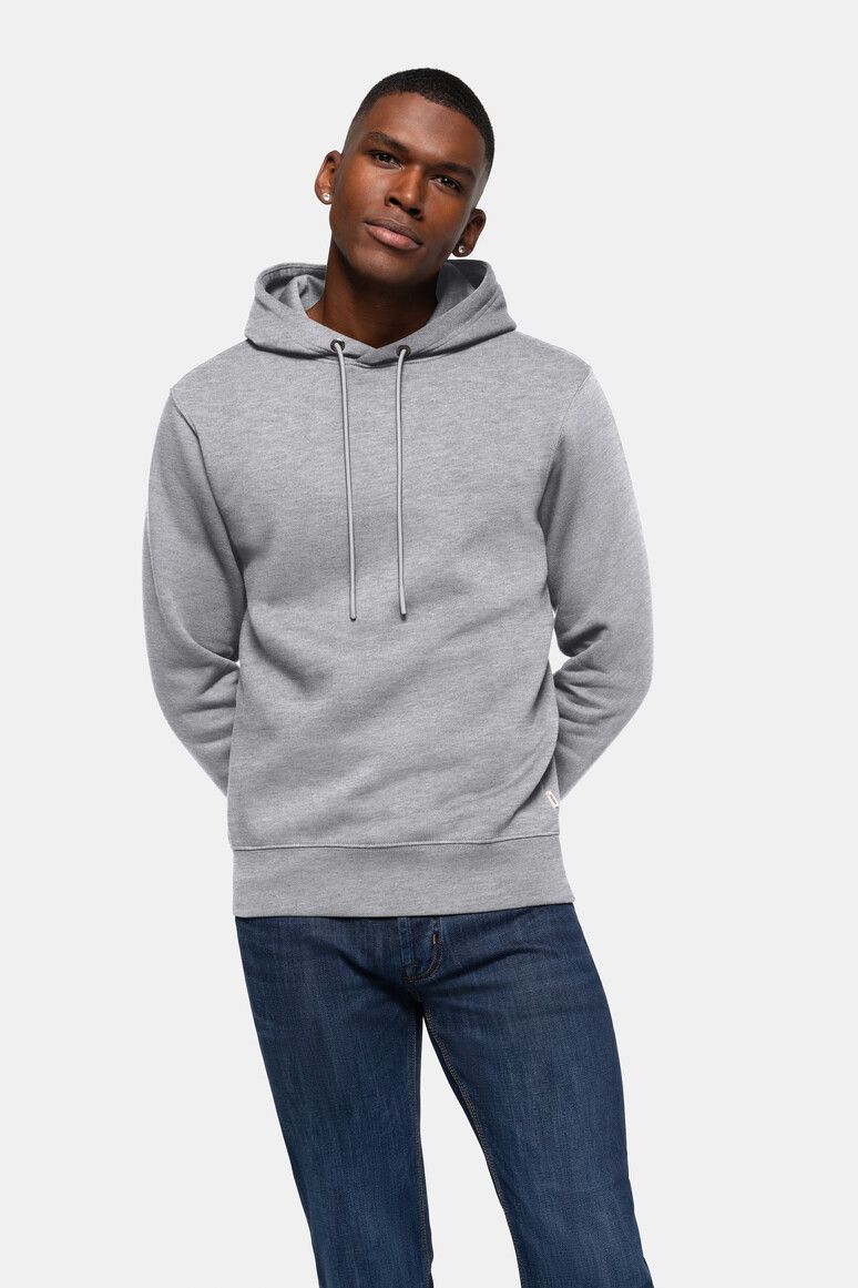 Mann in grauem Kapuzenpullover und Bluejeans, Hände hinter dem Rücken, blickt in die Kamera.