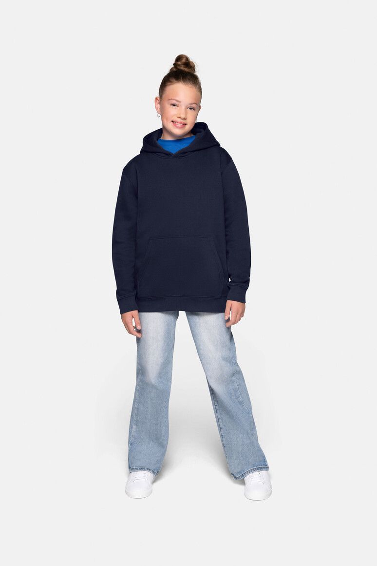 Mädchen mit dunkelblauem Kapuzenpullover, blauem Hemd, hell gewaschenen Jeans und weißen Turnschuhen steht auf weißem Hintergrund.