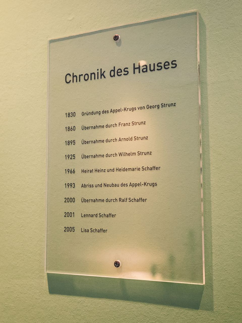 Eine klare Glastafel mit der Aufschrift „Chronik des Hauses“ und einer Liste von Daten und Ereignissen.