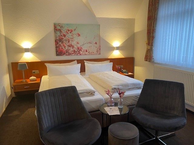Hotelzimmer mit zwei Betten, zwei grauen Stühlen und einem kleinen Tisch. Über den Betten hängt Blumenkunst.