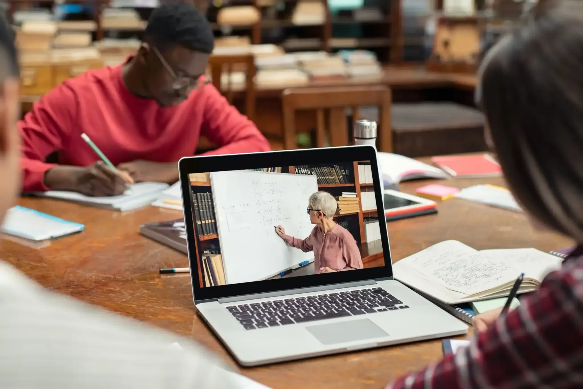 Los estudiantes en la biblioteca miran la pantalla de una computadora portátil que muestra a un profesor dando una lección.