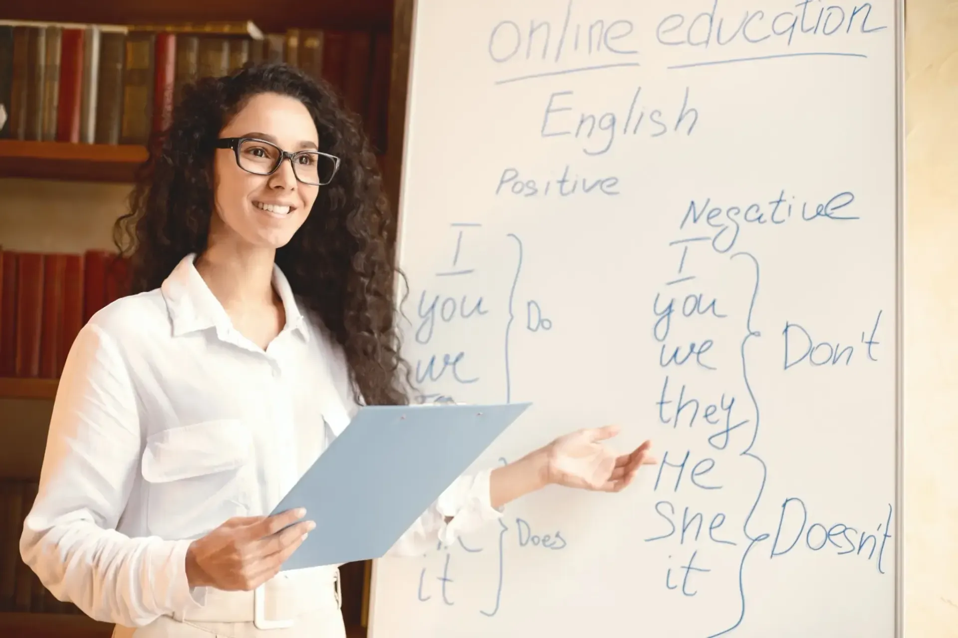 Mujer enseñando inglés, señalando una pizarra con notas gramaticales, sonriendo. Estantería al fondo.