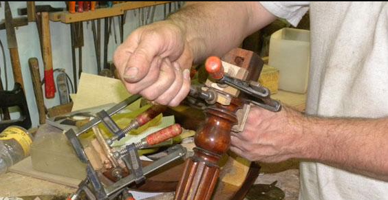 Dans un atelier, une personne utilise de petites pinces métalliques pour fixer des pièces de bois sur un pied de meuble sombre et orné.