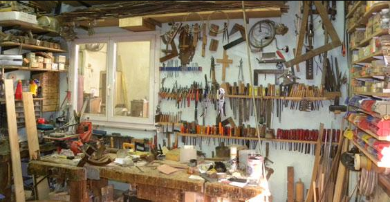 Un atelier de menuiserie encombré, avec un établi, divers outils à main accrochés aux murs et des étagères de rangement.