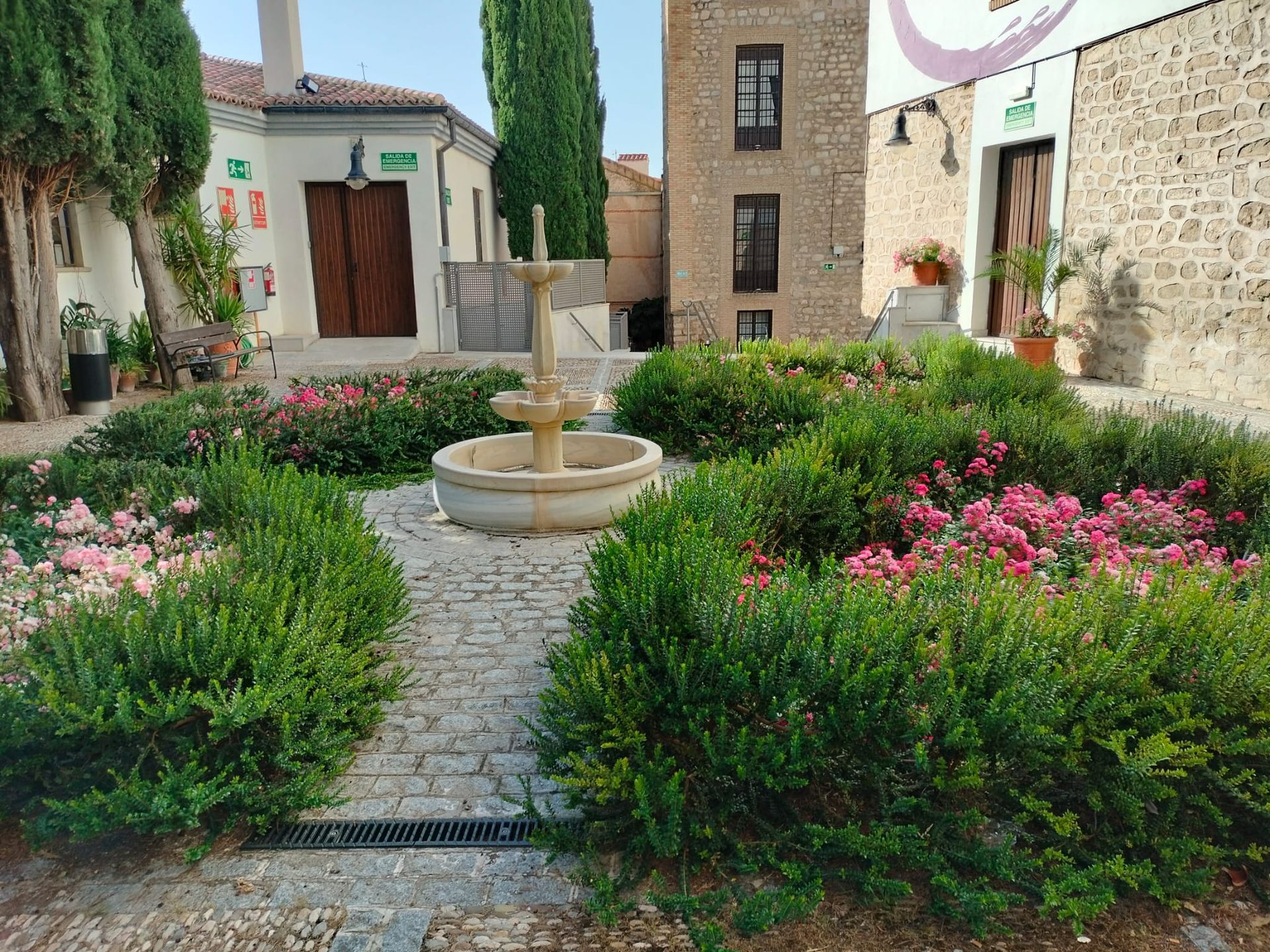 Un patio con una fuente de piedra rodeada de arbustos verdes, flores rosas y edificios bajo un cielo azul.