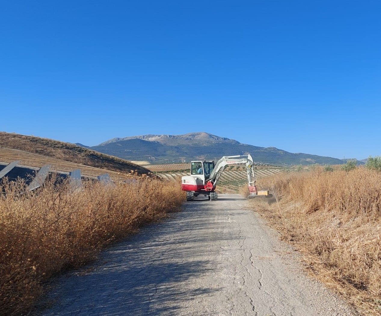 Una pequeña excavadora trabaja en un camino de grava en un campo con montañas y cielo azul de fondo.