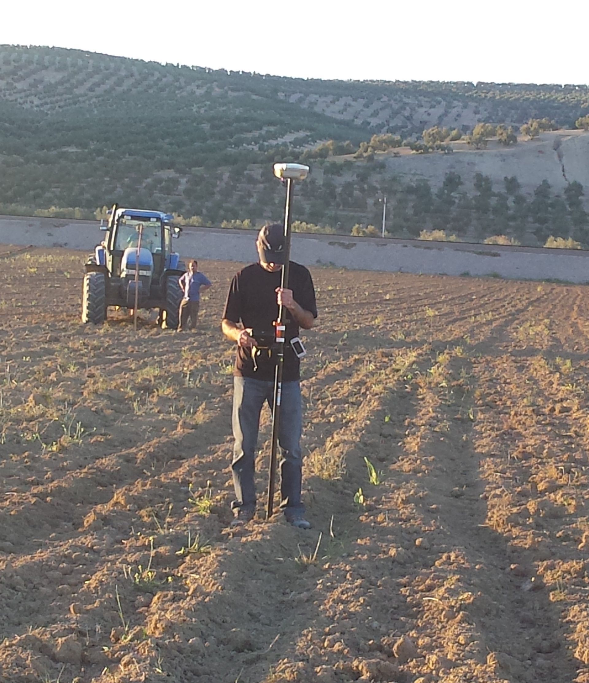 Hombre usando un GPS en un campo, con un tractor al fondo. El escenario es un campo arado, posiblemente agrícola.