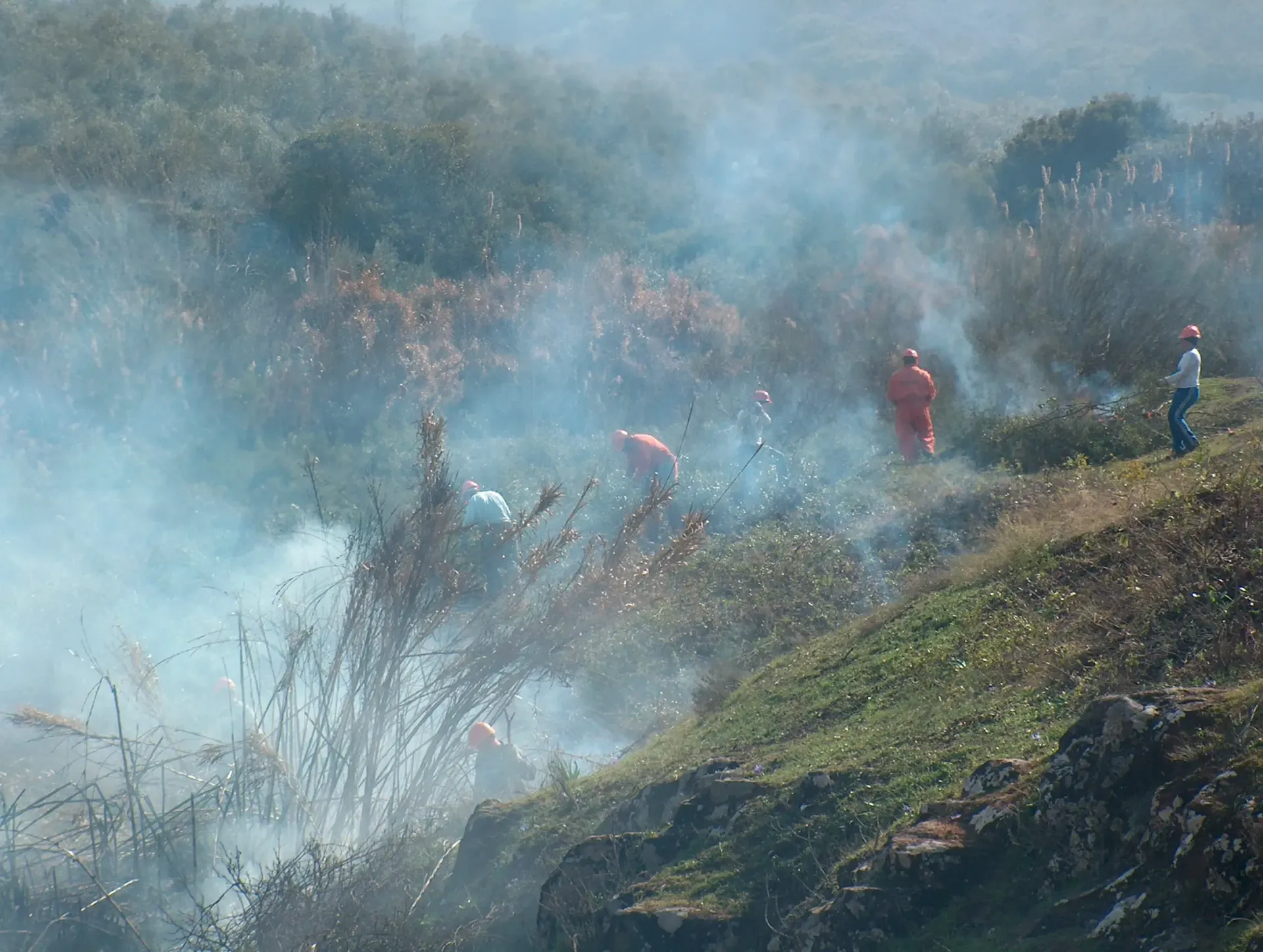 Bomberos con trajes naranjas luchan contra un incendio forestal en una ladera cubierta de hierba, rodeados de humo y escombros voladores.