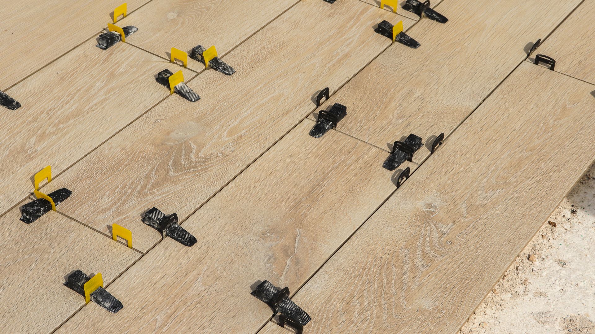 Gros plan sur la pose de carreaux beiges avec des clips de nivellement jaunes et de la colle noire disposées en diagonale.