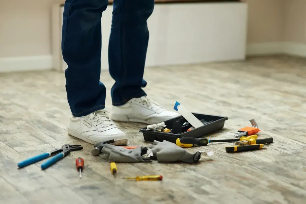 Persona de pie con herramientas esparcidas sobre un piso de madera, entorno interior.