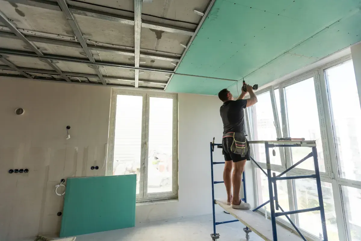 Un hombre instala paneles de yeso en el techo de una habitación con ventanas grandes, utilizando un andamio.