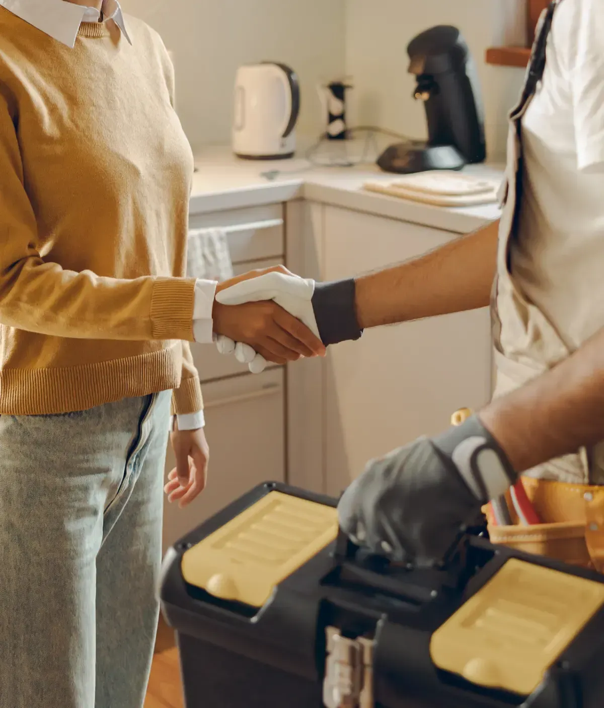 Mujer y reparador se dan la mano en una cocina, caja de herramientas en primer plano.