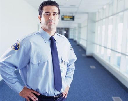 Un guardia de seguridad con uniforme azul claro y corbata está de pie con las manos en las caderas en un pasillo luminoso y moderno del aeropuerto.