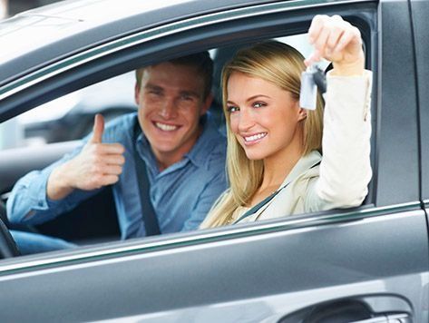 Un hombre sonriente levanta el pulgar y una mujer sostiene las llaves de un coche dentro de un vehículo estacionado.