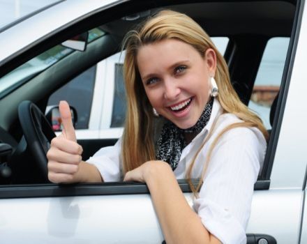 Una persona sonriente en un coche levanta el pulgar, vestida con una camisa blanca y una bufanda estampada.