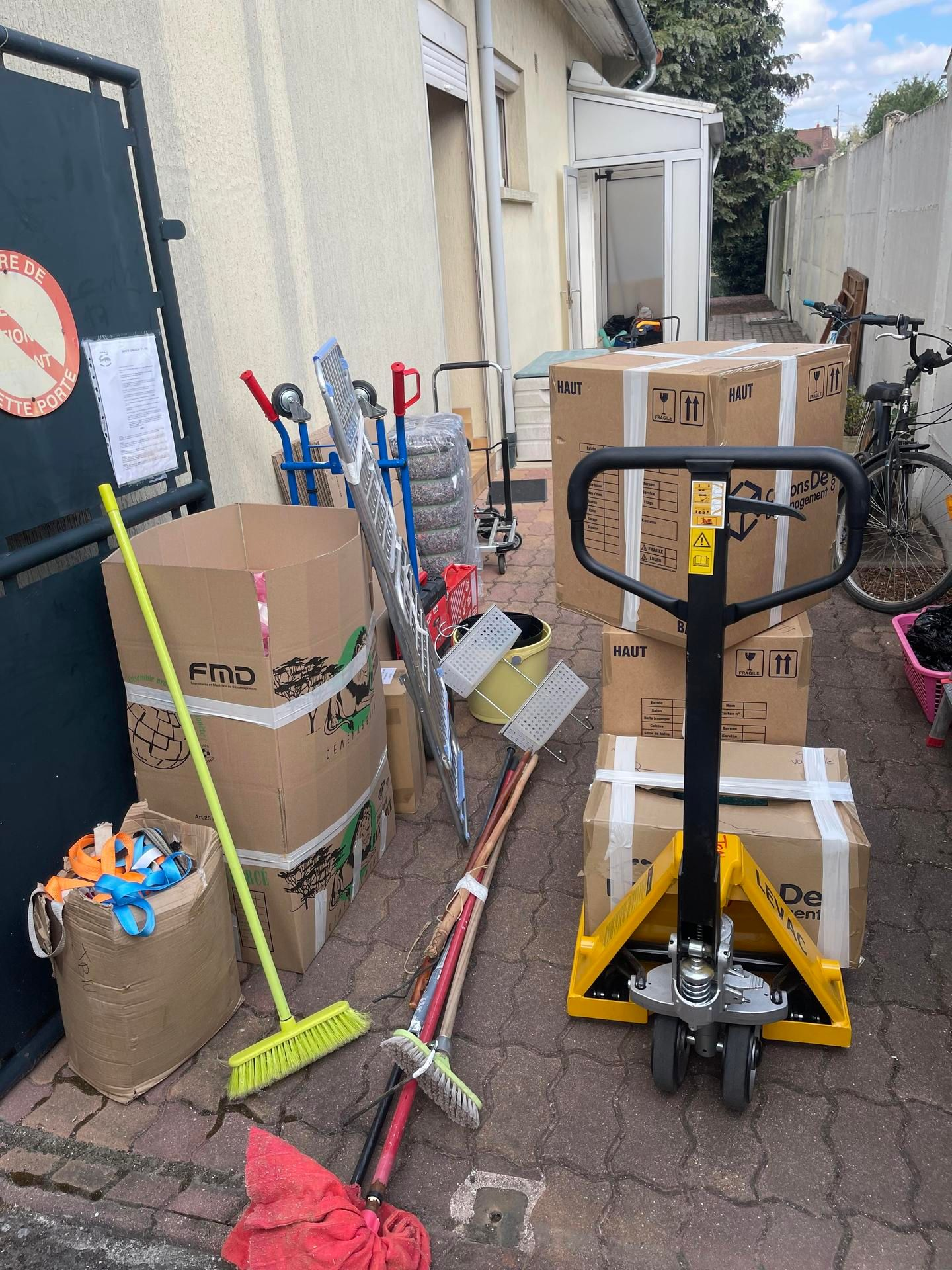Des cartons, des balais, des outils et un transpalette à l'extérieur d'un bâtiment.