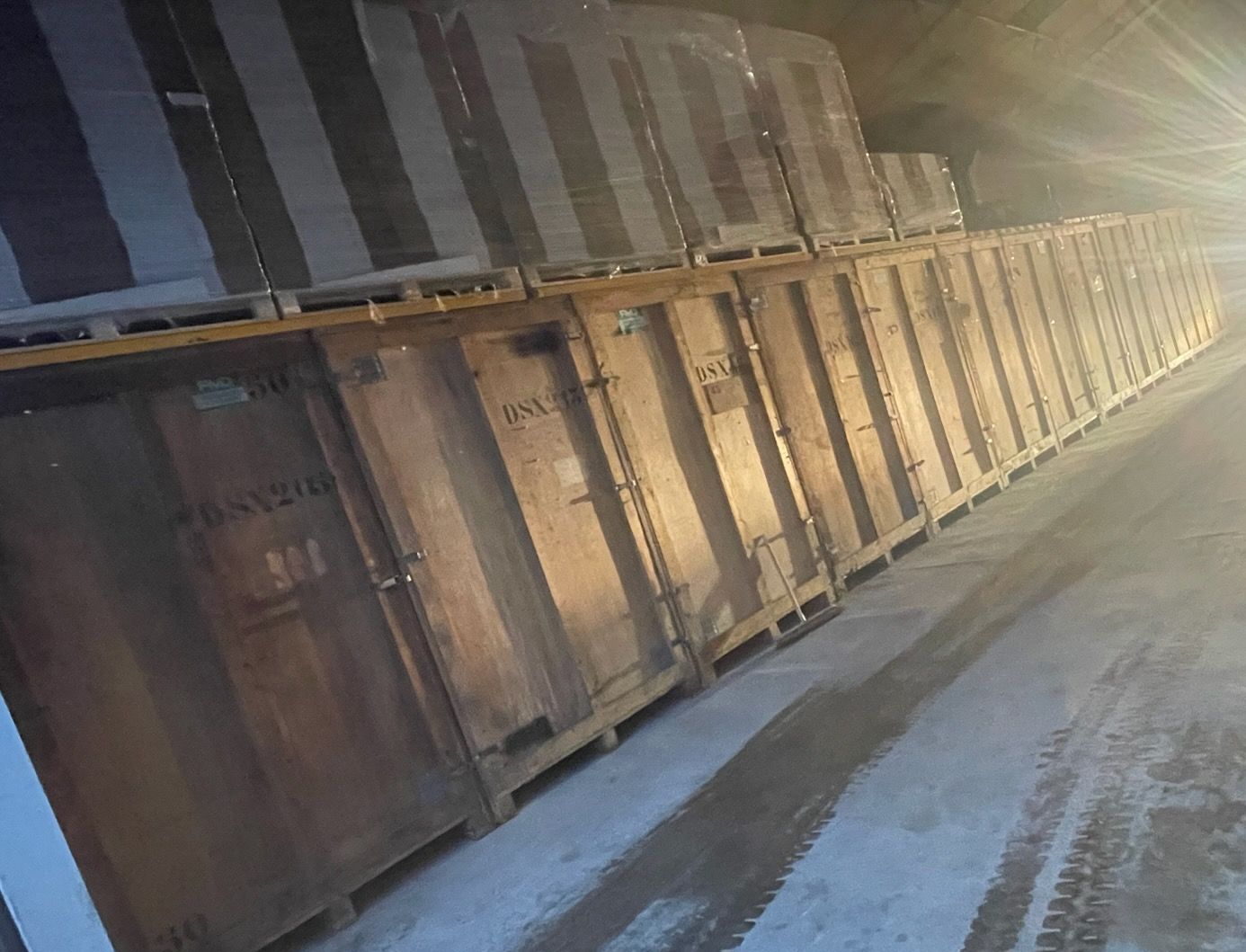 Des caisses en bois sont empilées dans un entrepôt. Des palettes de marchandises emballées sont empilées sur les caisses.