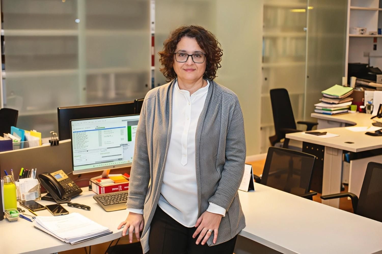 Mujer con gafas hablando por teléfono, sentada en un escritorio con una computadora.