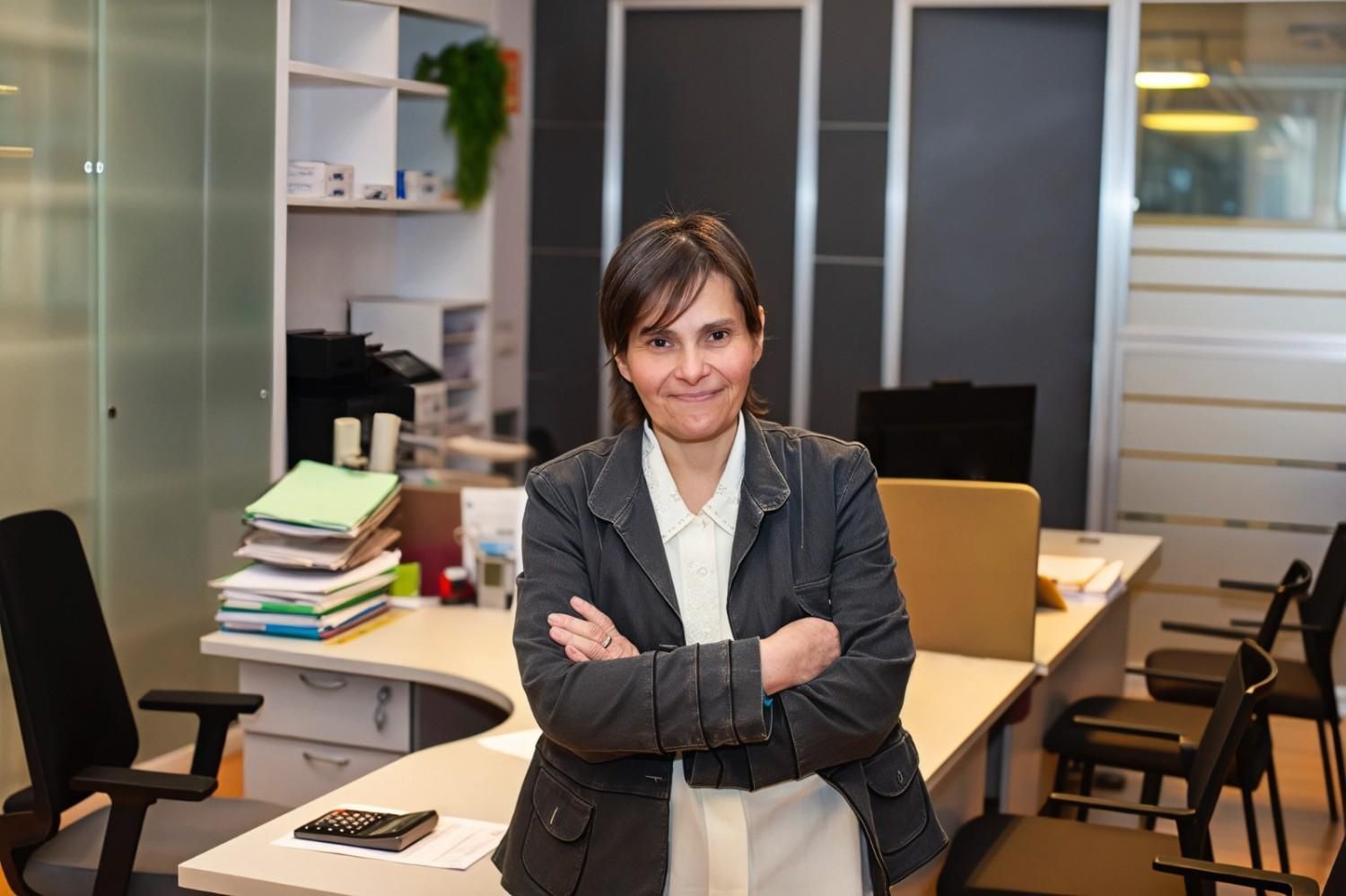 Mujer con pelo corto y gafas, trabajando en una computadora, con documentos en un escritorio.