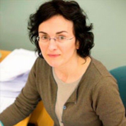 Mujer con gafas y suéter marrón, sonriendo levemente, sentada en el interior.