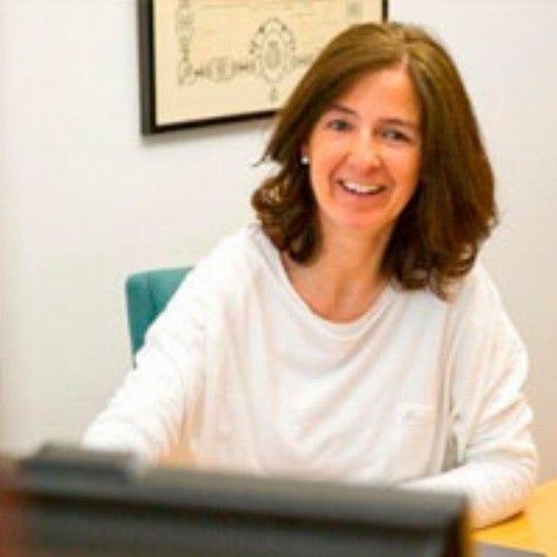Una mujer de cabello castaño sonríe, sentada en un escritorio de oficina. Lleva una camisa blanca de manga larga.