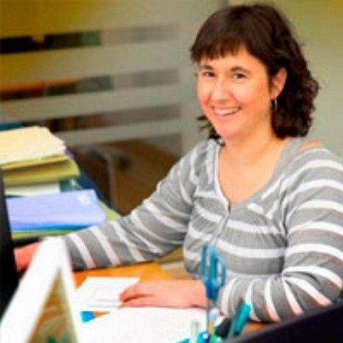 Mujer de cabello oscuro, sonriente, trabajando en un escritorio con papeles. Camisa a rayas, en un entorno de oficina.