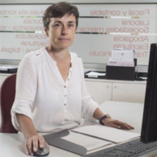 Mujer sentada en un escritorio usando una computadora. Lleva blusa blanca. En un entorno de oficina, con papeles visibles.