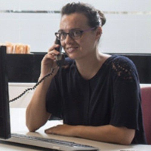 Mujer con gafas hablando por teléfono, sentada en un escritorio con una computadora.