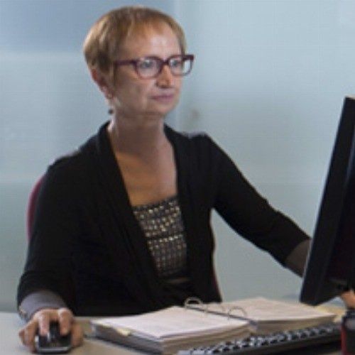 Mujer con pelo corto y gafas, trabajando en una computadora, con documentos en un escritorio.