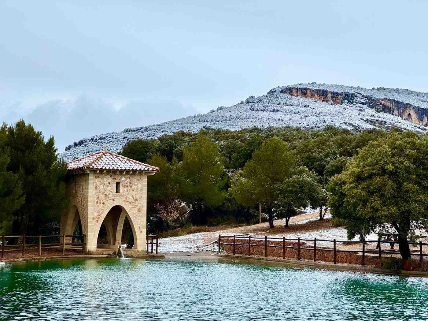Un cenador de piedra se alza junto a un estanque tranquilo de color turquesa, con una ligera capa de nieve.