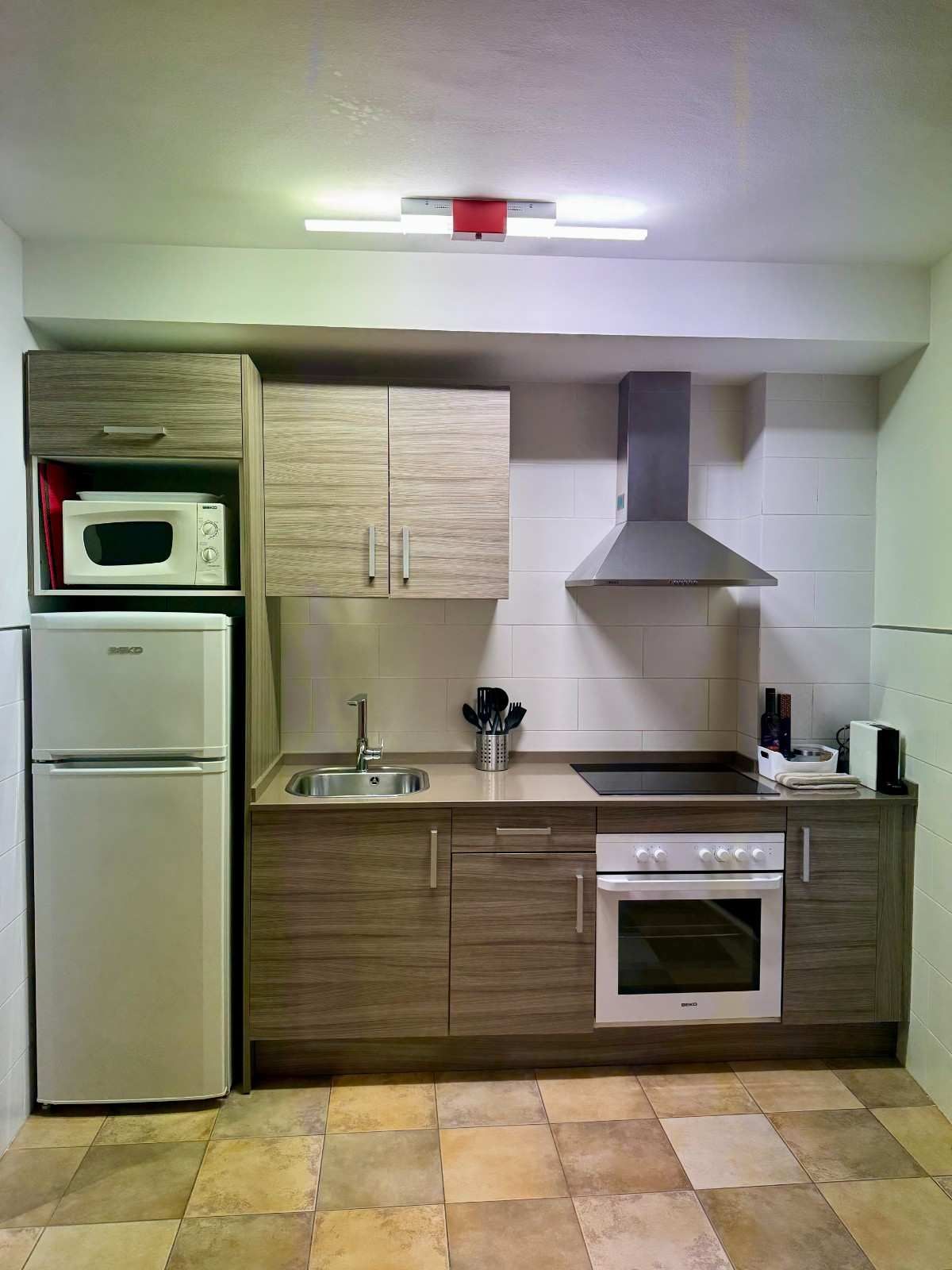 Una cocina compacta con armarios de madera de color marrón, un frigorífico blanco, microondas, fogones y campana extractora.