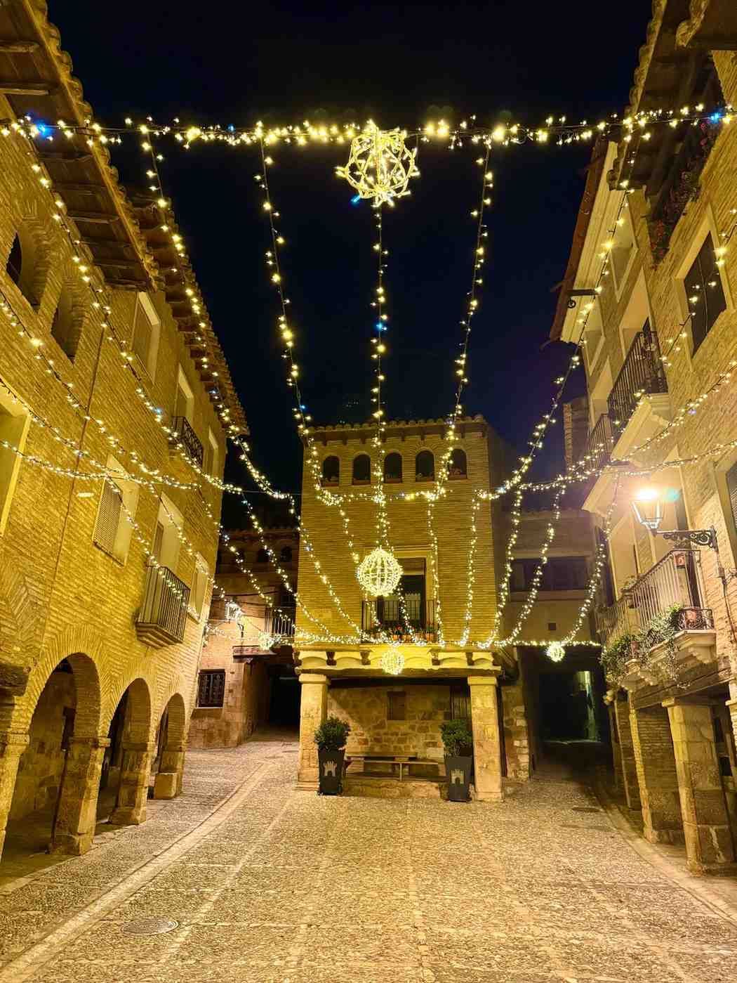 Una calle empedrada de noche, bordeada de edificios de piedra e iluminada por guirnaldas festivas de luces doradas colgantes.