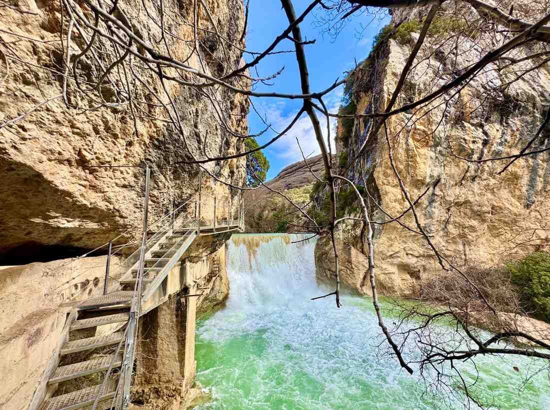 Una cascada de color turquesa fluye entre imponentes y escarpadas paredes de roca, con una escalera.