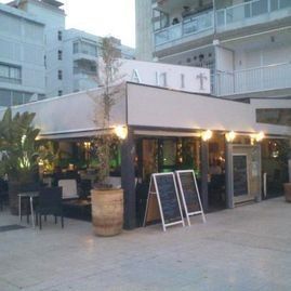 Cafetería al aire libre con sillas de mimbre y toldo junto a un edificio de apartamentos al atardecer.