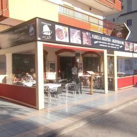 Cafetería al aire libre con asientos rojos y blancos en una soleada esquina, bajo toldos negros.