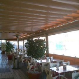 Terraza cubierta al aire libre con mesas, sillas, plantas en macetas y vistas luminosas al mar.
