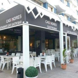Cafetería al aire libre con toldo negro, mesas y sillas blancas y plantas en macetas en la acera.